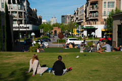 The lawn at a mixed-use development in Alpharetta, Ga. with people sitting in grass, cars going by store fronts, on May 9. (Kevin D. Liles/For the Washington Post)